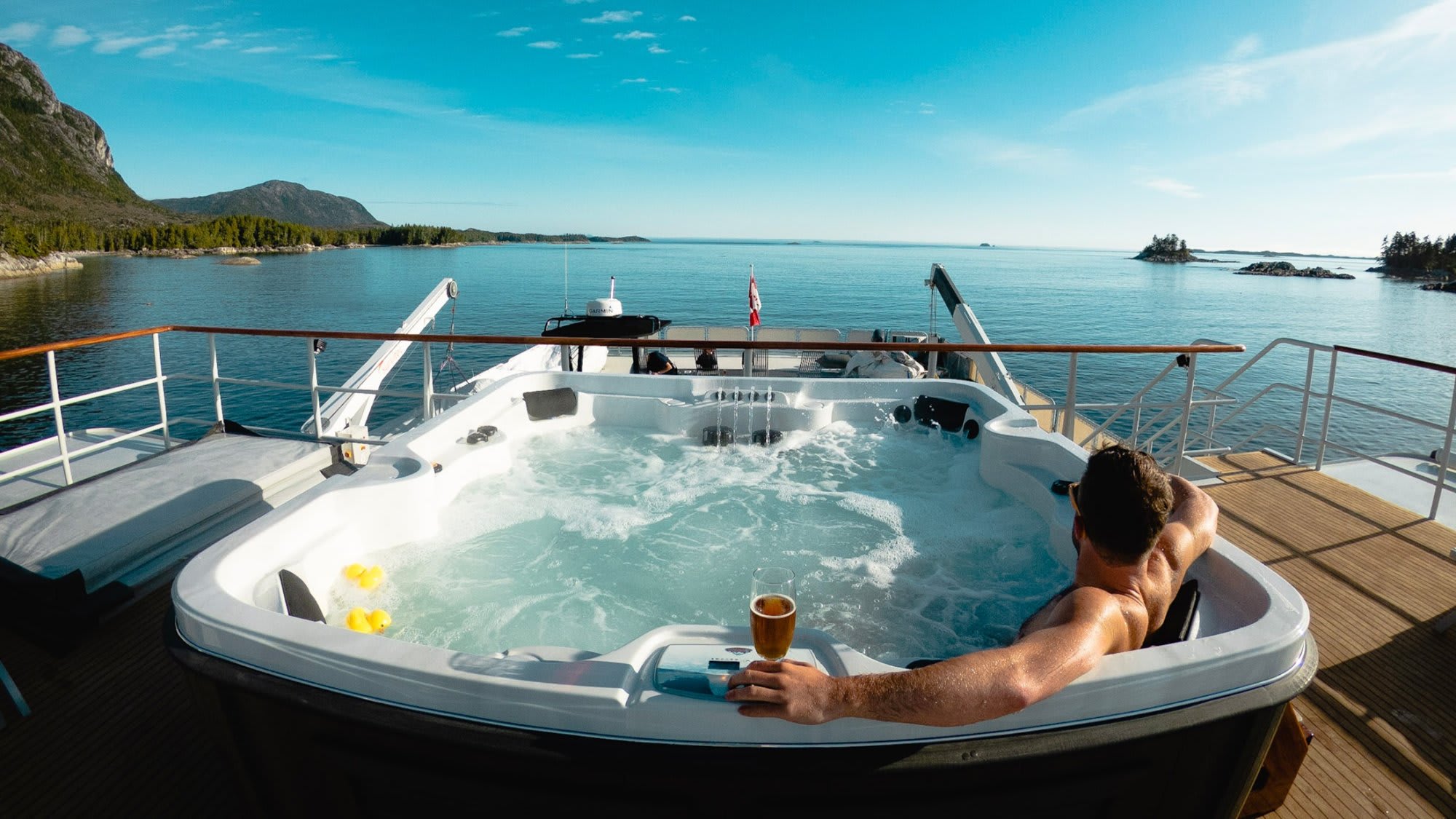 Luxury Hot Tub on Yacht in Lake A person relaxes in a large, bubbling hot tub on the deck of a luxury yacht, holding a beverage while enjoying panoramic views of a pristine turquoise lake surrounded by forested mountains and rocky shores.