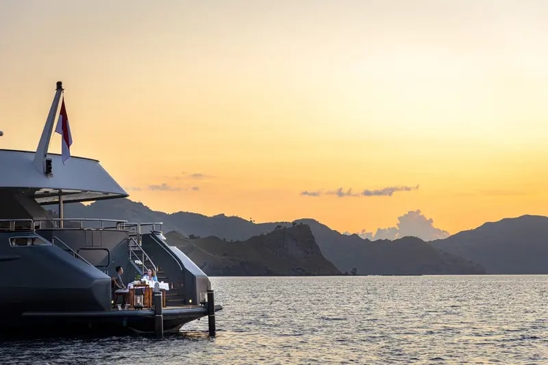 A modern luxury yacht tender is moored at sunset with dramatic golden-orange skies reflecting off calm waters, with mountainous tropical islands silhouetted in the background.
