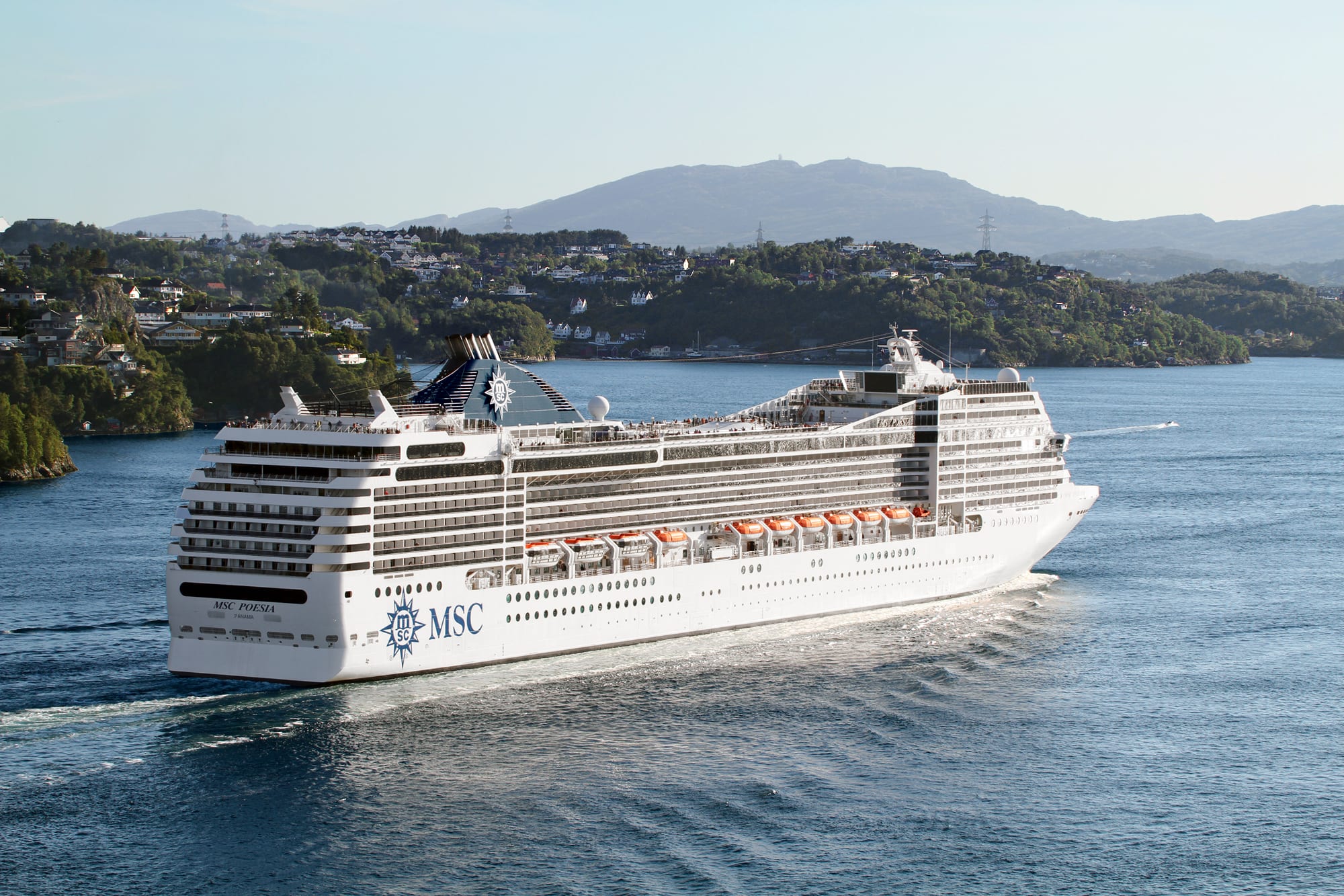 A large white MSC cruise ship sailing through a picturesque bay with wooded shorelines