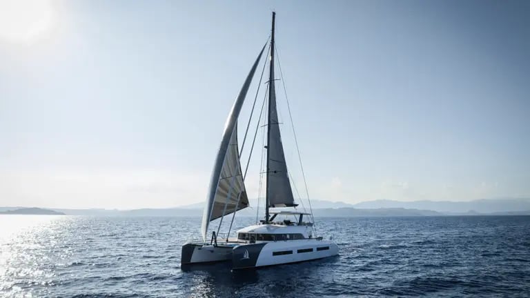 A sleek white and gray catamaran sailing with fully raised sails on a calm, blue ocean with distant