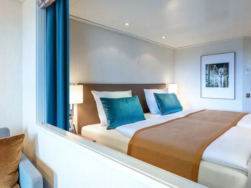 Modern Cruise Ship Cabin with Ocean View A contemporary cruise ship stateroom featuring a comfortable double bed with teal accent pillows, warm wood furnishings, and a large picture window with ocean views.