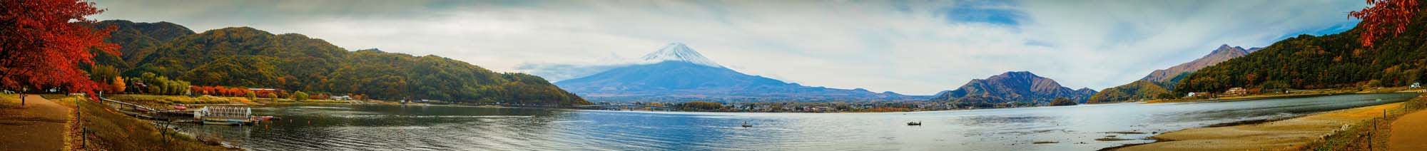 A panoramic view of Mount Fuji with a serene lake in the foreground, surrounded by mountains with vibrant autumn foliage in red and green colors.