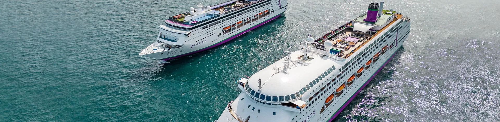 Multiple Cruise Ships Docked at Port A large modern cruise ship named Ambassador, featuring white hull with distinctive purple and orange accent bands, docked at a tropical port with clear blue waters. The vessel displays multiple decks, communication satellites, and lifeboats typical of contemporary ocean liner design.