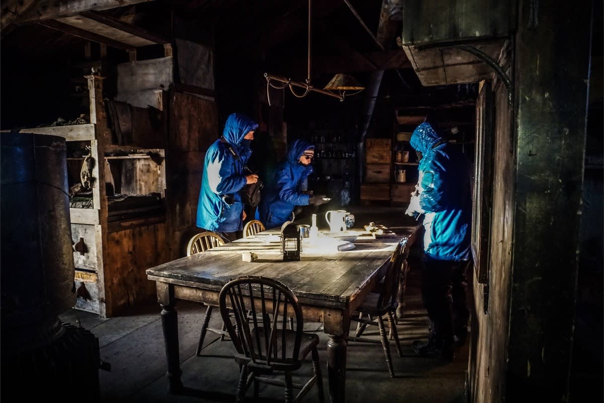 Three people in bright blue jackets gather around a weathered wooden table in a dimly-lit, rustic