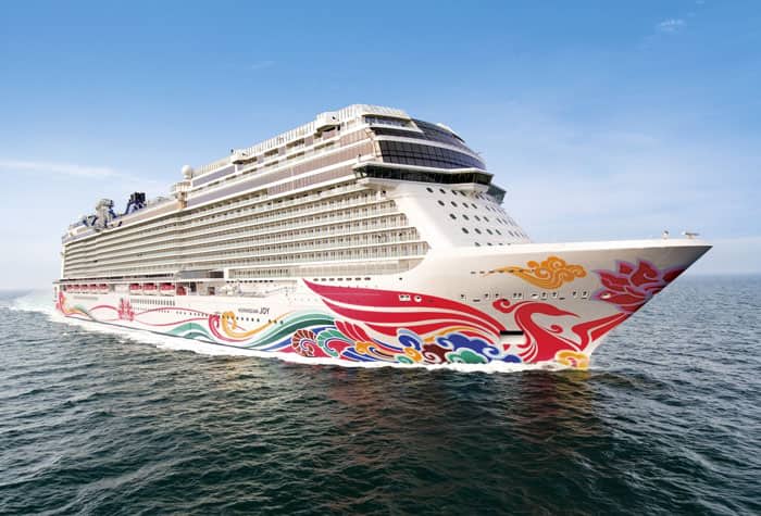 Norwegian Cruise Ship at Sea A large, colorful Norwegian cruise ship sailing on open water, featuring a vibrant red, yellow, and blue hull design. The ship appears modern and sleek against the blue sky and ocean.