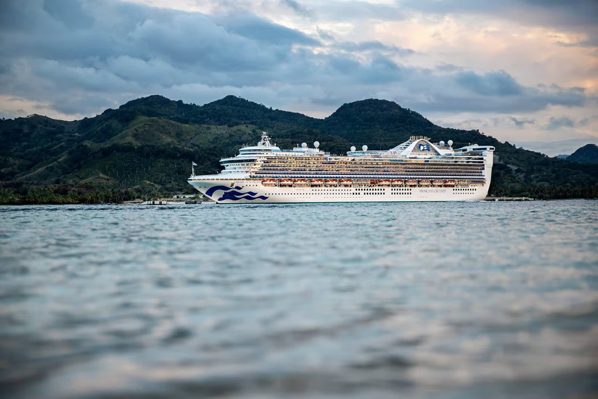 A large white Princess Cruises ship anchored in a scenic bay with lush green mountains in