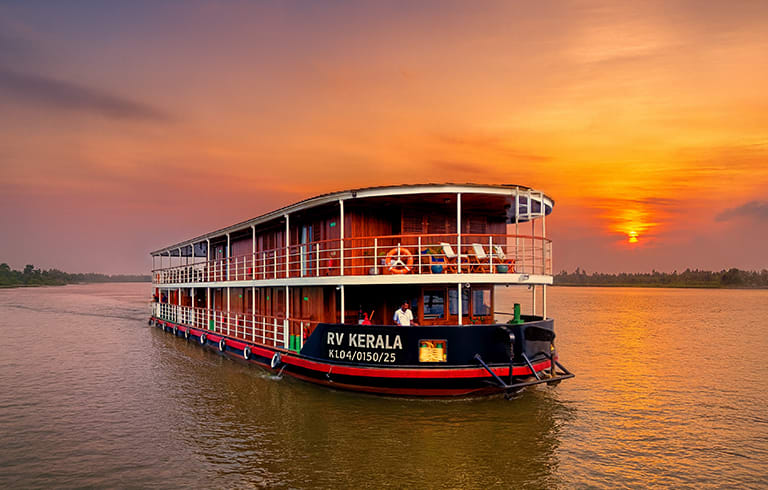 A multi-deck river cruise boat named RV Kerala sailing on a tranquil river during a vibrant orange