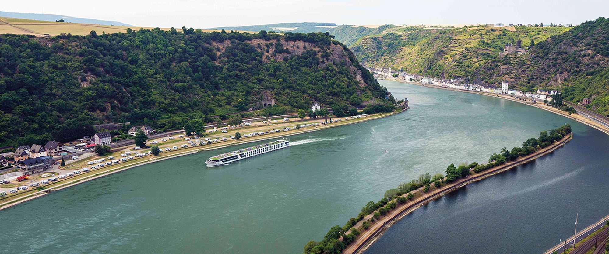 Rhine River Cruise Through Scenic Valley A scenic aerial view of a river cruise ship navigating through the dramatic Rhine Gorge, with steep forested cliffs, a charming riverside village, and lush green hillsides.