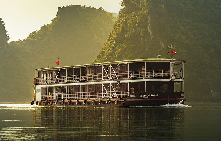 A multi-level river cruise boat navigating through a scenic landscape with lush green mountains and misty morning light. The vessel appears to be a river cruise ship sailing on a tranquil waterway.