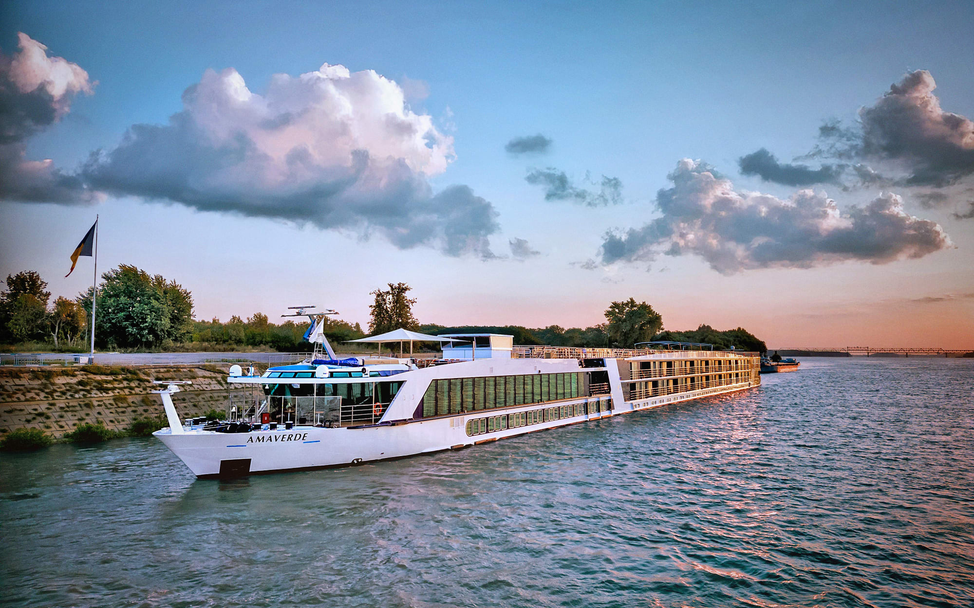 River Cruise Ship Amaverde at Sunset A white river cruise ship named Amaverde docked along a river at sunset, with dramatic cloudy skies and a tree-lined shore in the background. The vessel reflects on the calm water with soft golden and blue tones.