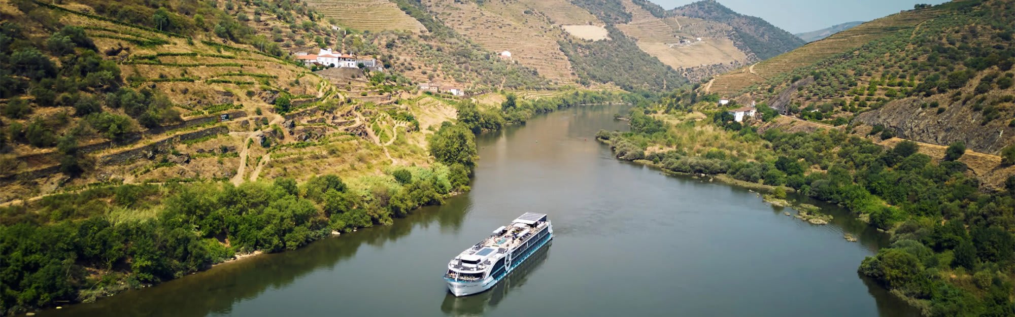 A modern river cruise ship navigates through the scenic Douro River valley, surrounded by terraced vineyards on steep hillsides and traditional villages perched on the landscape.