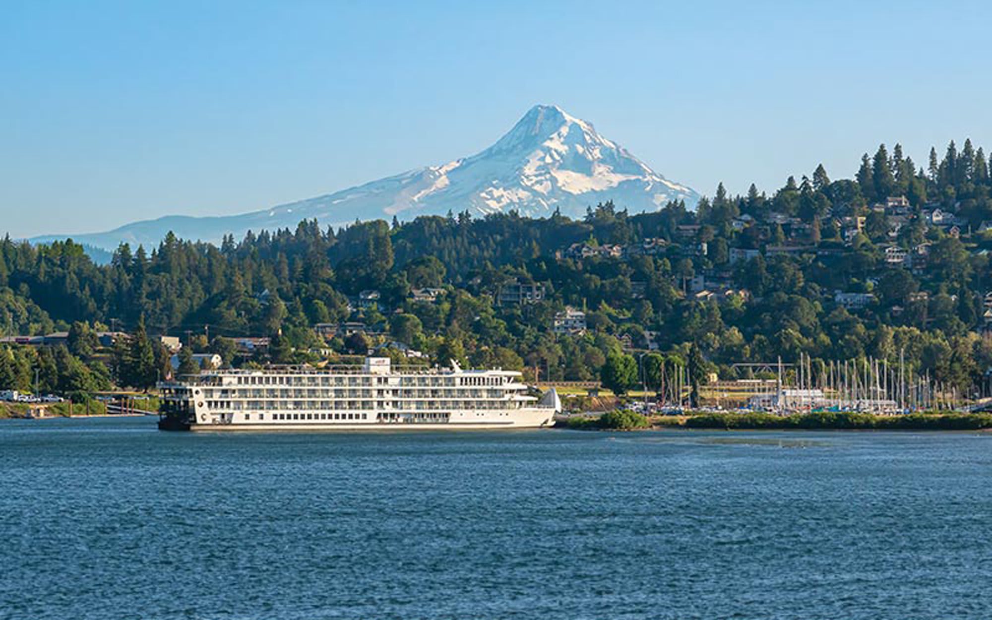 Riverboat Cruise at Mount Hood, Oregon A multi-deck white riverboat navigates calm blue waters with snow-capped Mount Hood dominating the background, surrounded by dense evergreen forests and a hillside community nestled among the trees in the Pacific Northwest.