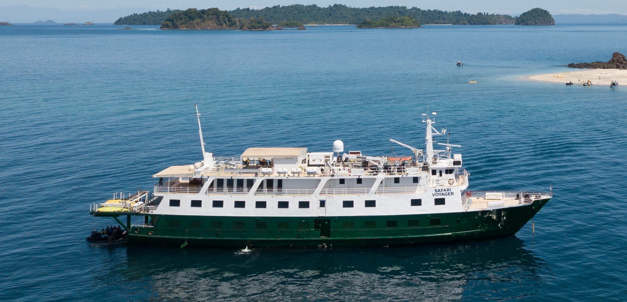 A green and white expedition cruise ship anchored in calm blue waters with lush tropical islands in the background. The Safari Voyager appears to be a small adventure cruise vessel positioned near a sandy beach and scenic archipelago.