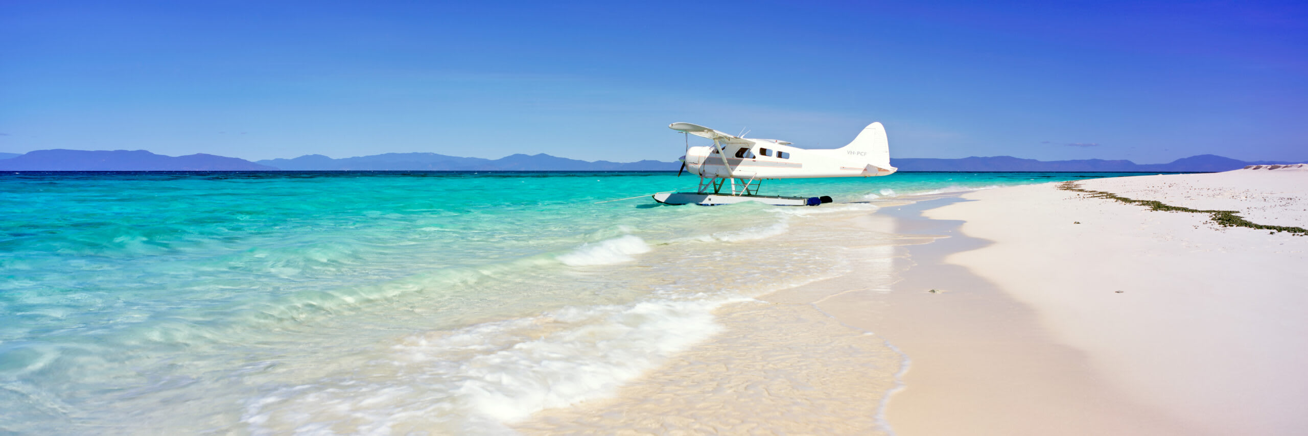 Seaplane on Pristine Tropical Beach A white twin-engine seaplane is moored in the crystal-clear turquoise waters of a pristine tropical beach, with powdery white sand and calm shallow waters stretching to the shoreline. Mountains are visible across the horizon under a clear blue sky, suggesting a remote island destination perfect for scenic flights and water-based tourism.