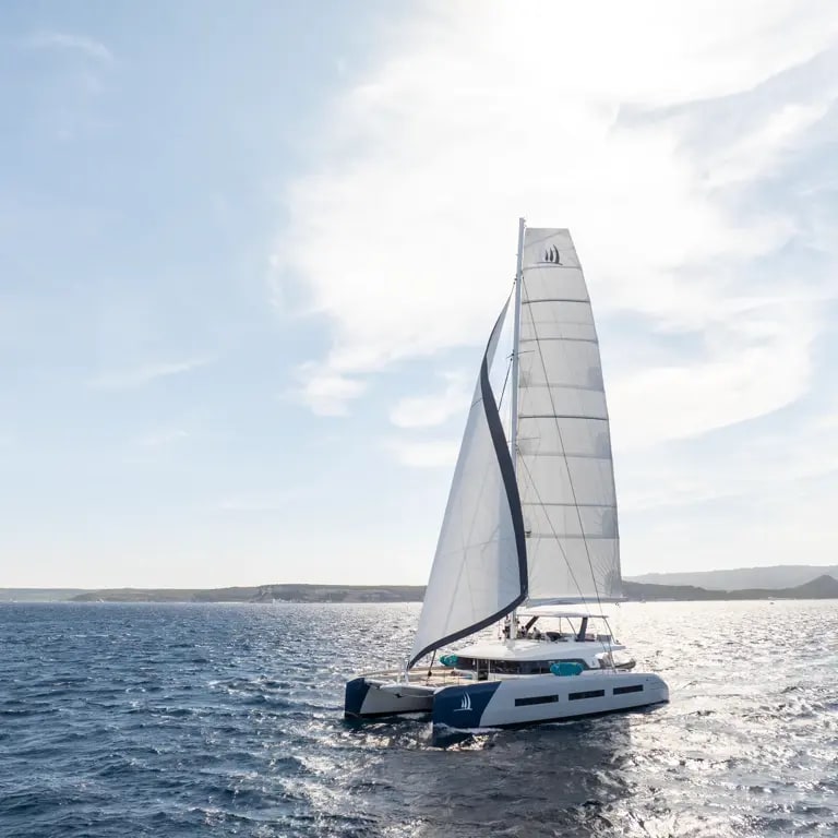 A modern white catamaran with fully raised sails navigating through bright blue ocean waters
