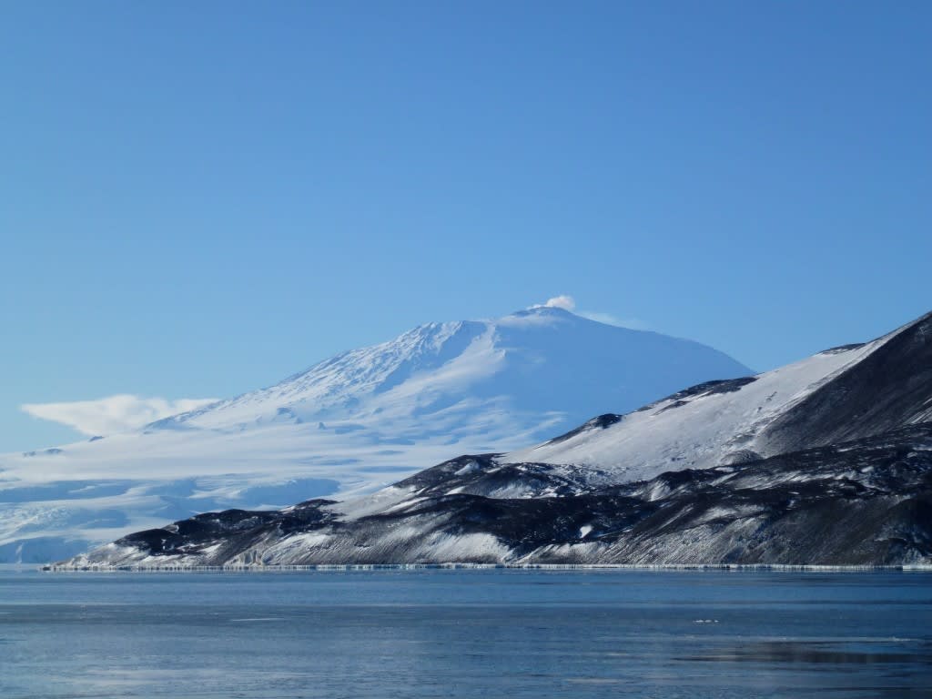 A majestic snow-covered volcanic peak rises dramatically above a frozen lake with dark volcanic