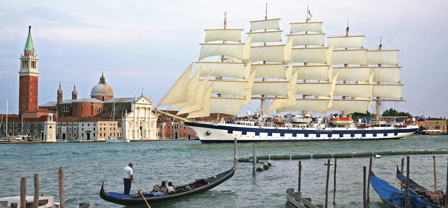 A magnificent multi-masted sailing vessel with full white sails anchored in the Venice lagoon, with historic Italian architecture and landmarks in the background.