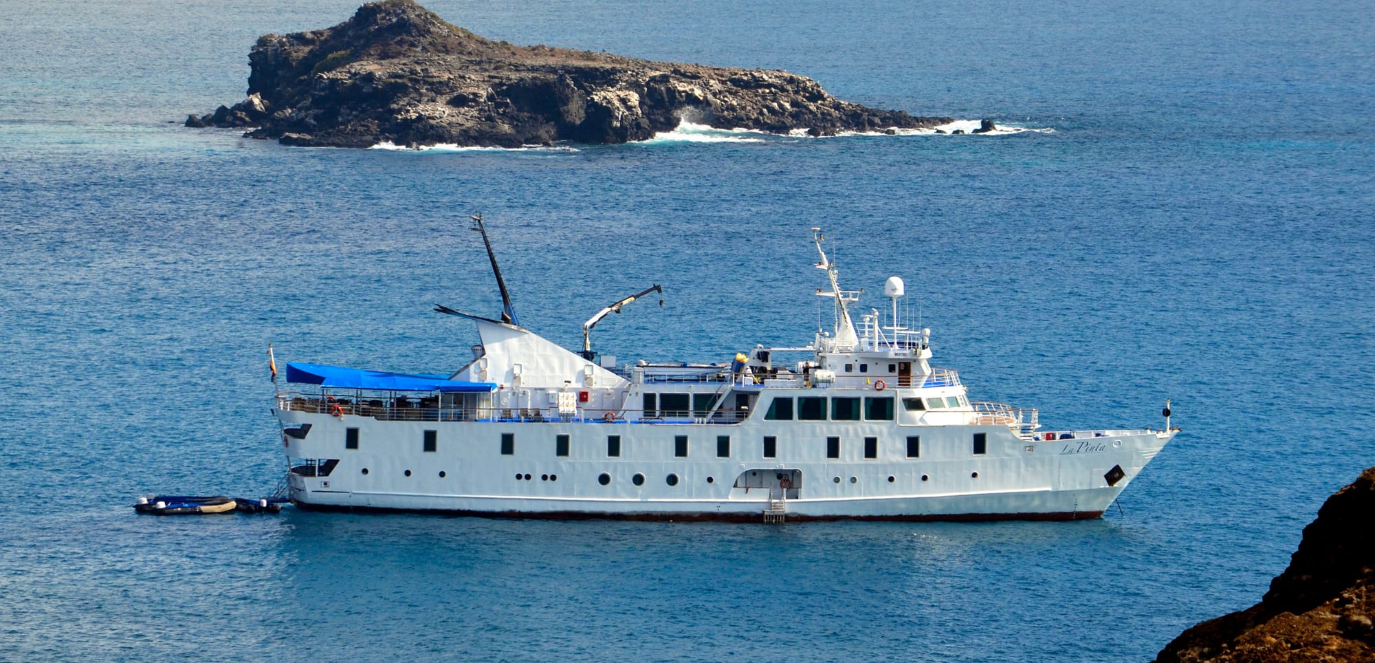A white expedition-style cruise vessel anchored near a rocky, volcanic island with blue ocean waters surrounding it. The ship appears to be a small, specialized cruise boat with multiple decks and navigation equipment.