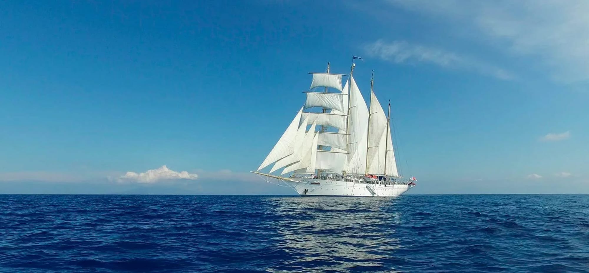 A classic three-masted tall ship with fully unfurled white sails cruising on a pristine blue ocean under a clear sky. The elegant vessel appears to be a modern sailing yacht or training ship.