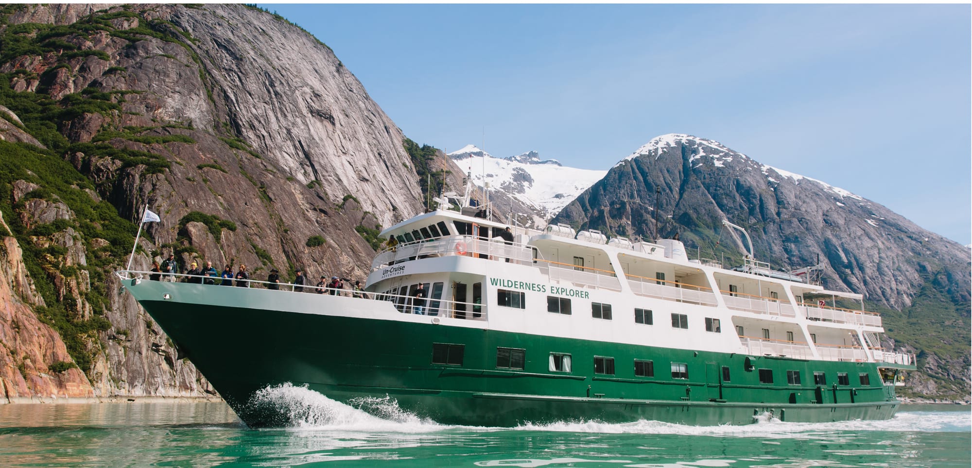 A green and white expedition cruise ship navigating through a scenic Alaskan fjord with dramatic