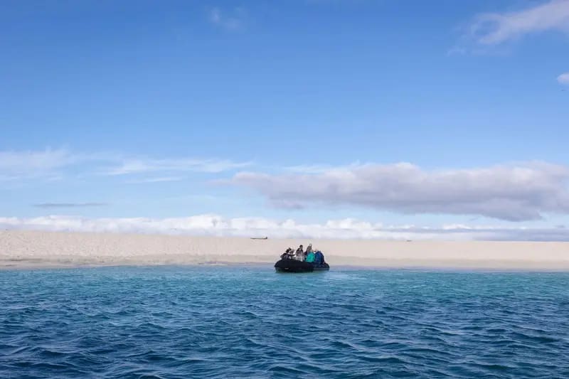 A small Zodiac landing craft with passengers approaches across turquoise Arctic waters, with a snow-covered mountainous coastline visible in the background under a partly cloudy sky.