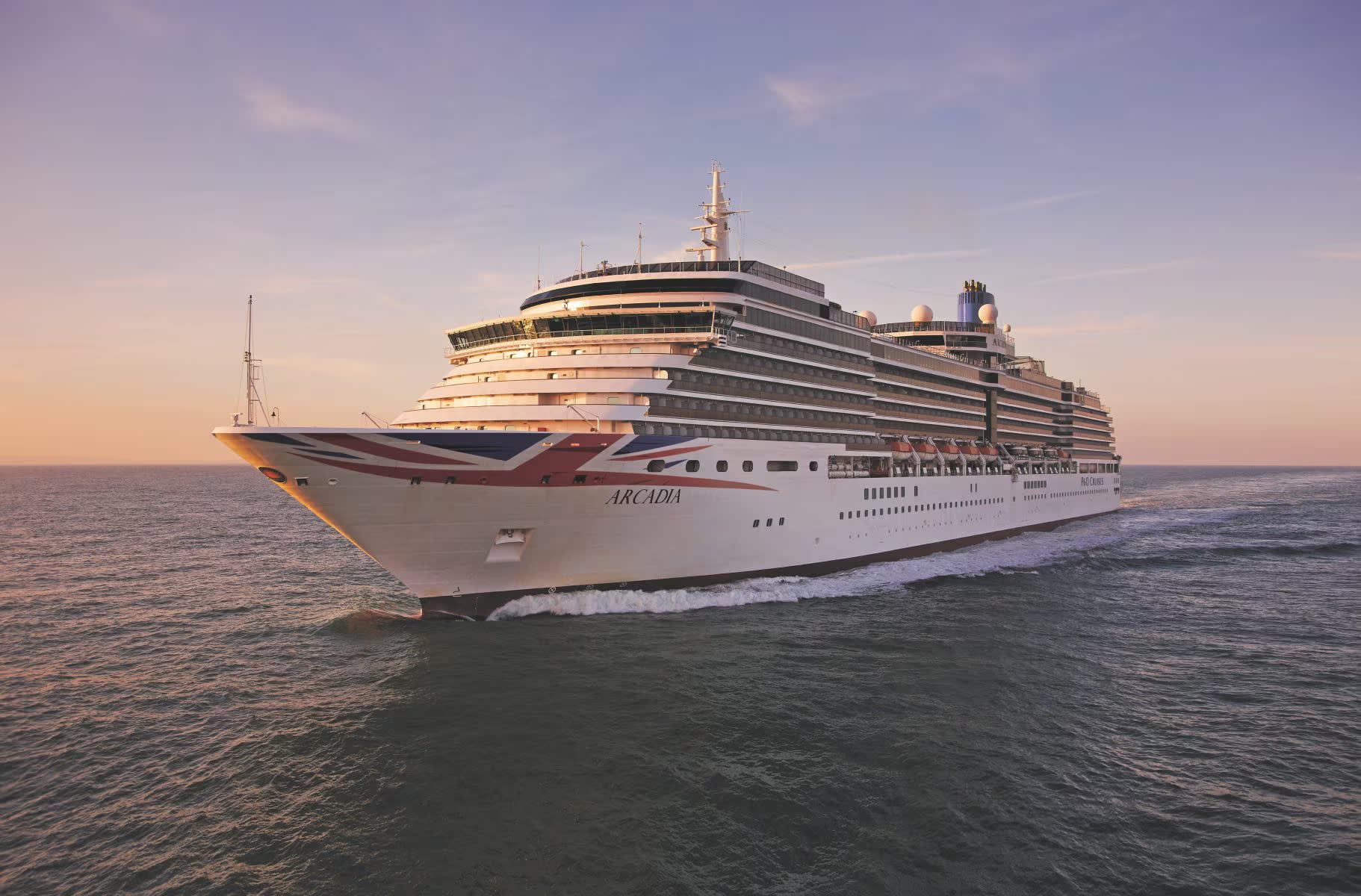 arcadia-at-sea-sunset-pocjpg1580833548223image18221200high The P&O Arcadia, a large modern cruise ship, is pictured in shallow coastal waters at golden hour. The vessel displays the distinctive P&O livery with Union Jack design on its hull, with multiple decks visible against a serene twilight sky and calm sea.