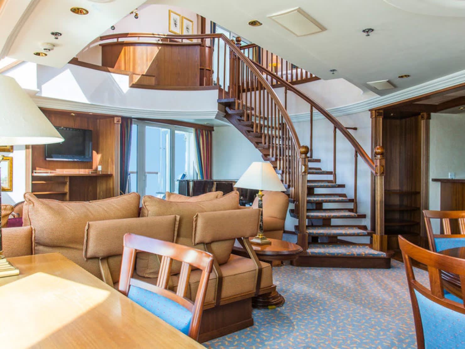 aurora-penthouse-29-18749704-post-2019-refit-18749704jpgimage14801110high A luxurious penthouse suite aboard a cruise ship featuring a dramatic spiral staircase with wooden railings, elegant furnishings in warm tones, and a two-story open layout.