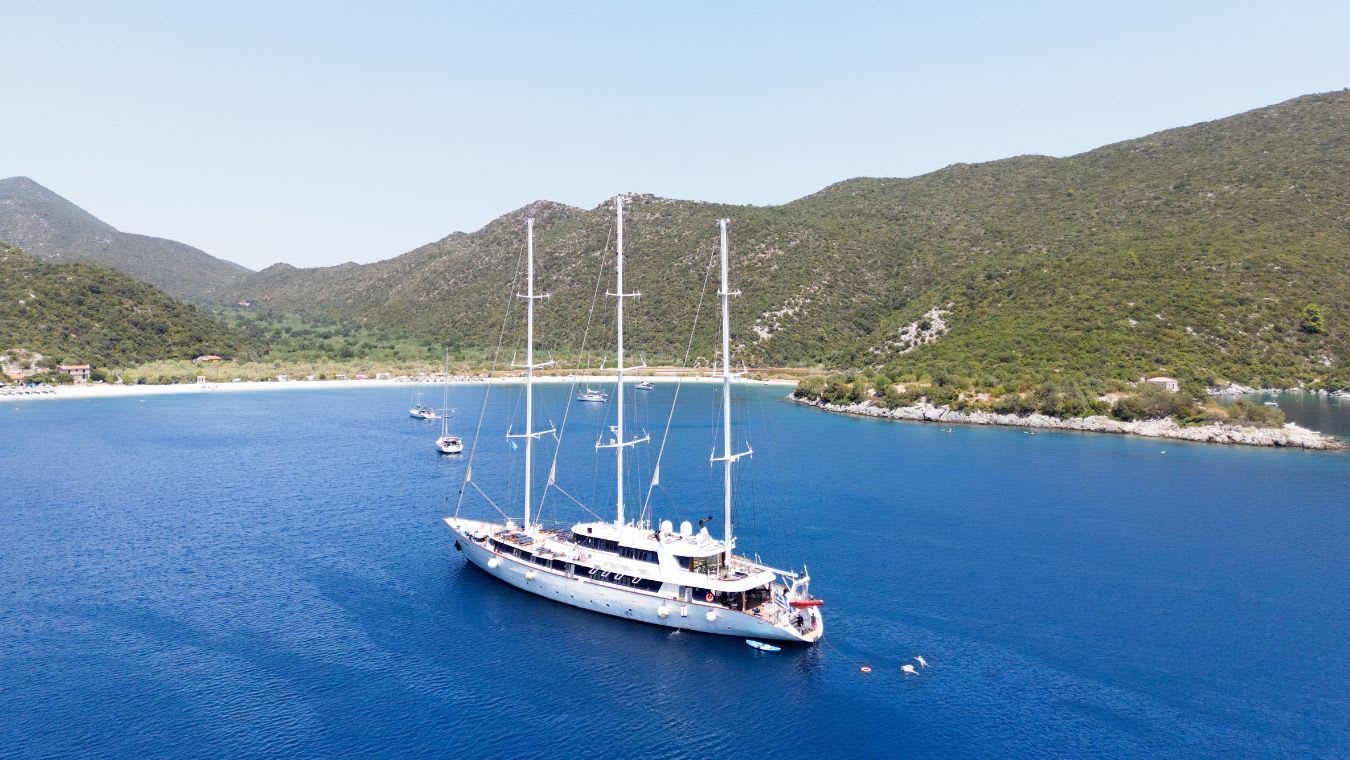 A stunning white multi-masted gulet sailing yacht anchors in crystal-clear turquoise waters of a picturesque Mediterranean bay, surrounded by verdant pine-covered mountains and a charming coastal village.