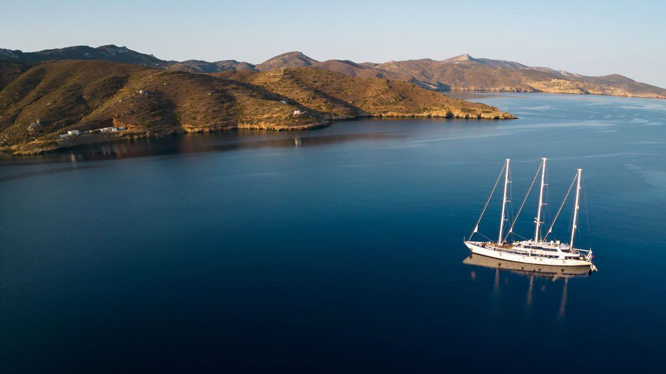 A stunning aerial view of a classic three-masted sailing vessel anchored in crystalline blue waters, with a scenic Mediterranean coastline featuring golden-brown hills and scattered white buildings.