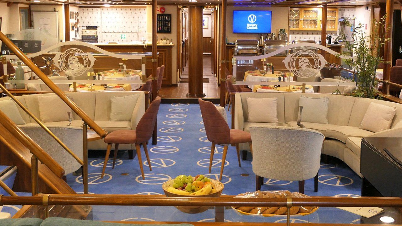 A sophisticated panorama lounge aboard a cruise ship featuring cream-colored seating arranged around dining tables, a blue patterned carpet with geometric designs, wooden architectural elements, and warm ambient lighting.