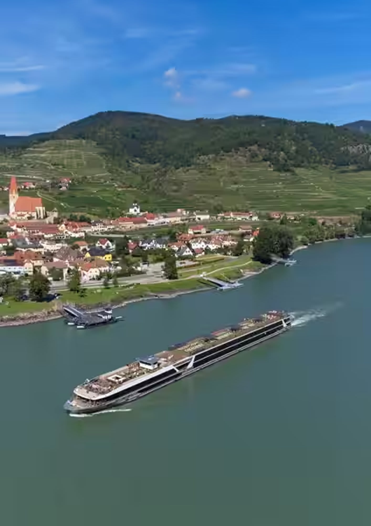 A modern river cruise ship navigates through a picturesque European river valley, passing a charming riverside town with a distinctive church steeple, terraced vineyards on rolling hills, and traditional architecture.