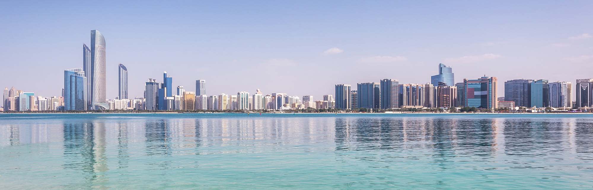 Abu Dhabi Skyline and Waterfront A panoramic view of Abu Dhabi's modern skyline reflecting in calm turquoise waters, showcasing numerous high-rise buildings and skyscrapers along the Persian Gulf coastline under clear blue skies.