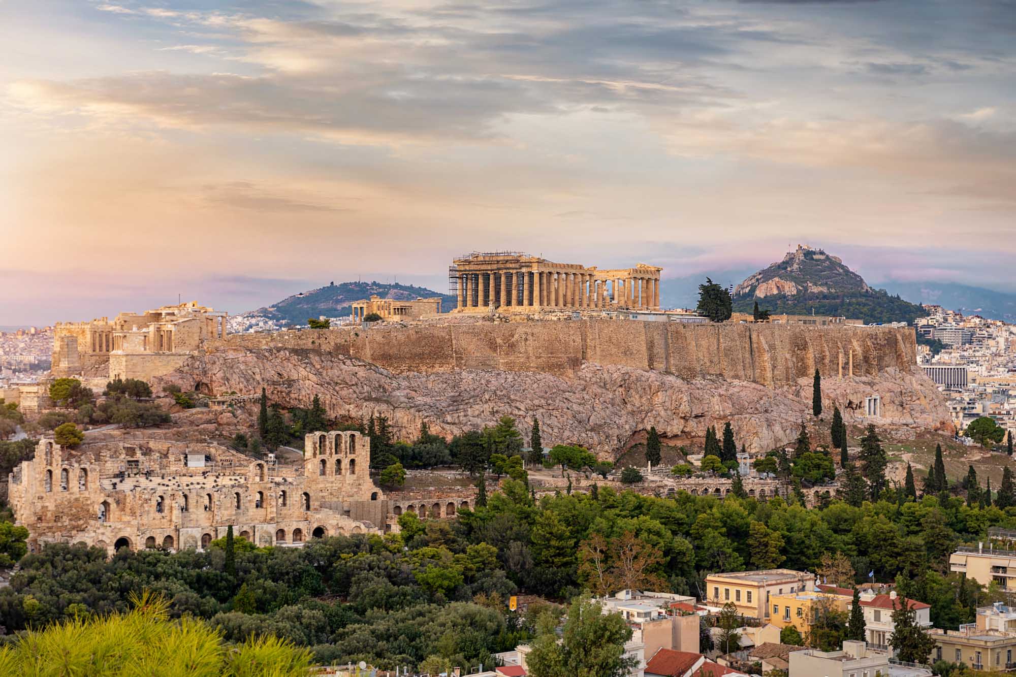 A stunning panoramic view of the Acropolis in Athens, Greece, showcasing the iconic Parthenon temple complex illuminated by golden sunset light atop a rocky outcrop, with the historic Plaka district, Lycabettus Hill, and modern Athens sprawling below under a dramatic cloudy sky.