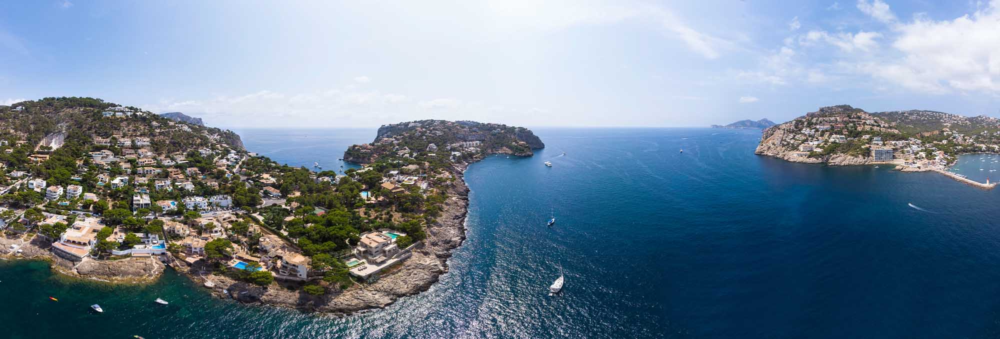 A stunning aerial photograph of Port d'Andratx in Mallorca, Balearic Islands, showcasing dramatic rocky coves, crystal-clear turquoise waters, luxury Mediterranean villas nestled on hillsides, and numerous moored yachts.