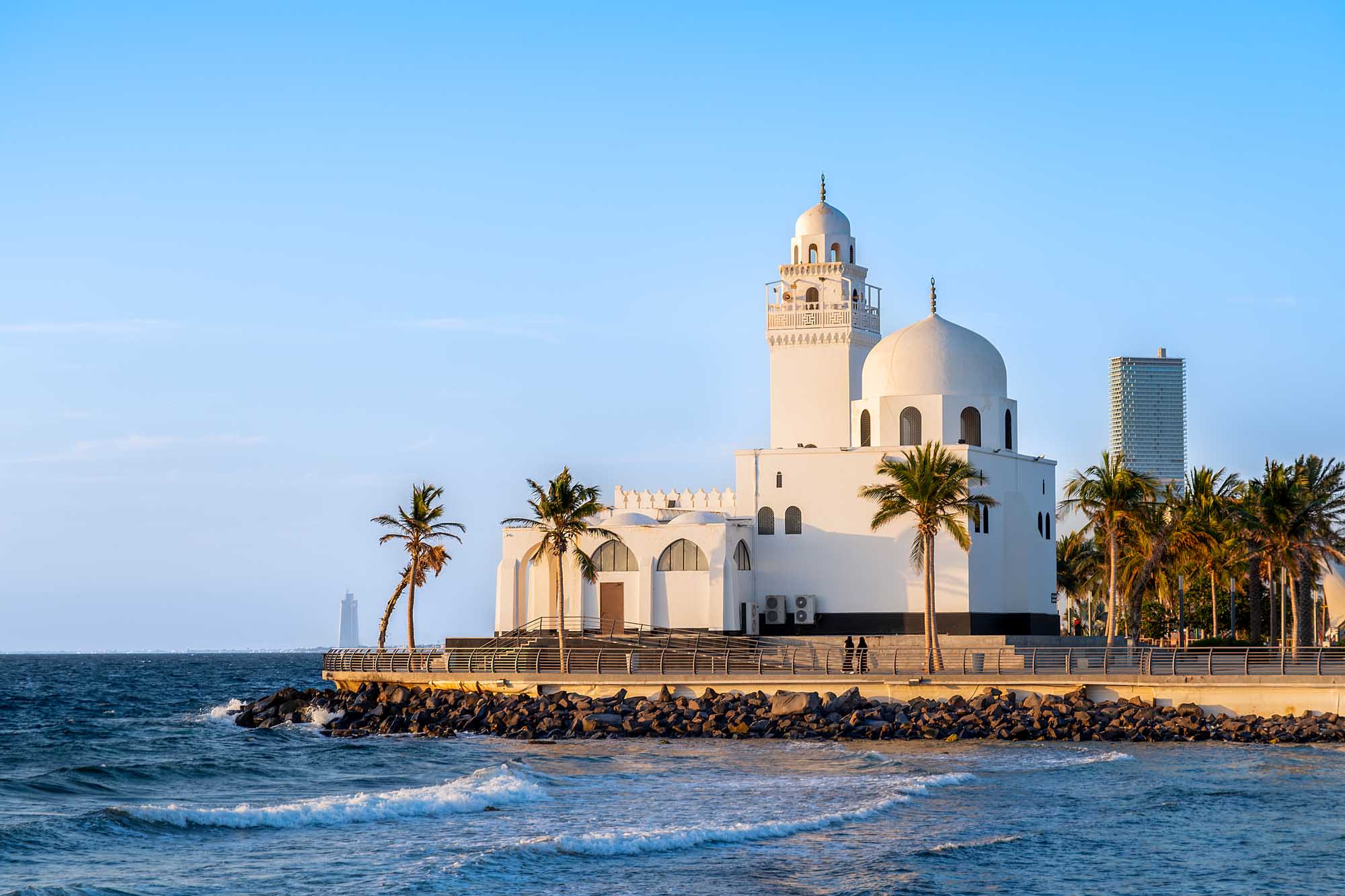 A striking white mosque with a large dome and minaret situated on the Jeddah waterfront, surrounded by palm trees and facing the Red Sea. The modern architectural landmark features traditional Islamic design elements and serves as a prominent symbol of contemporary Jeddah.