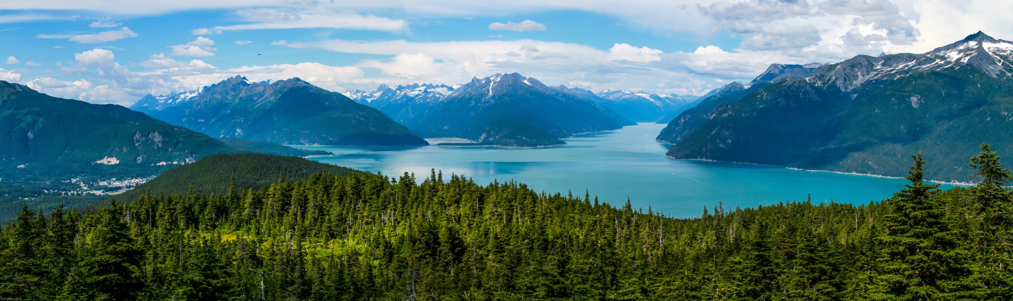A breathtaking panoramic view of a pristine turquoise alpine lake surrounded by dense coniferous forests and dramatic snow-capped mountain peaks.