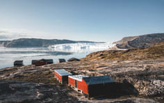 Arctic Cabin with Icebergs and Glacier A rustic orange-red cabin sits on a rocky Arctic shore overlooking a pristine fjord filled with icebergs and glaciers in the distance. The stark, barren landscape and massive ice formations exemplify the raw beauty of Arctic exploration and adventure travel.