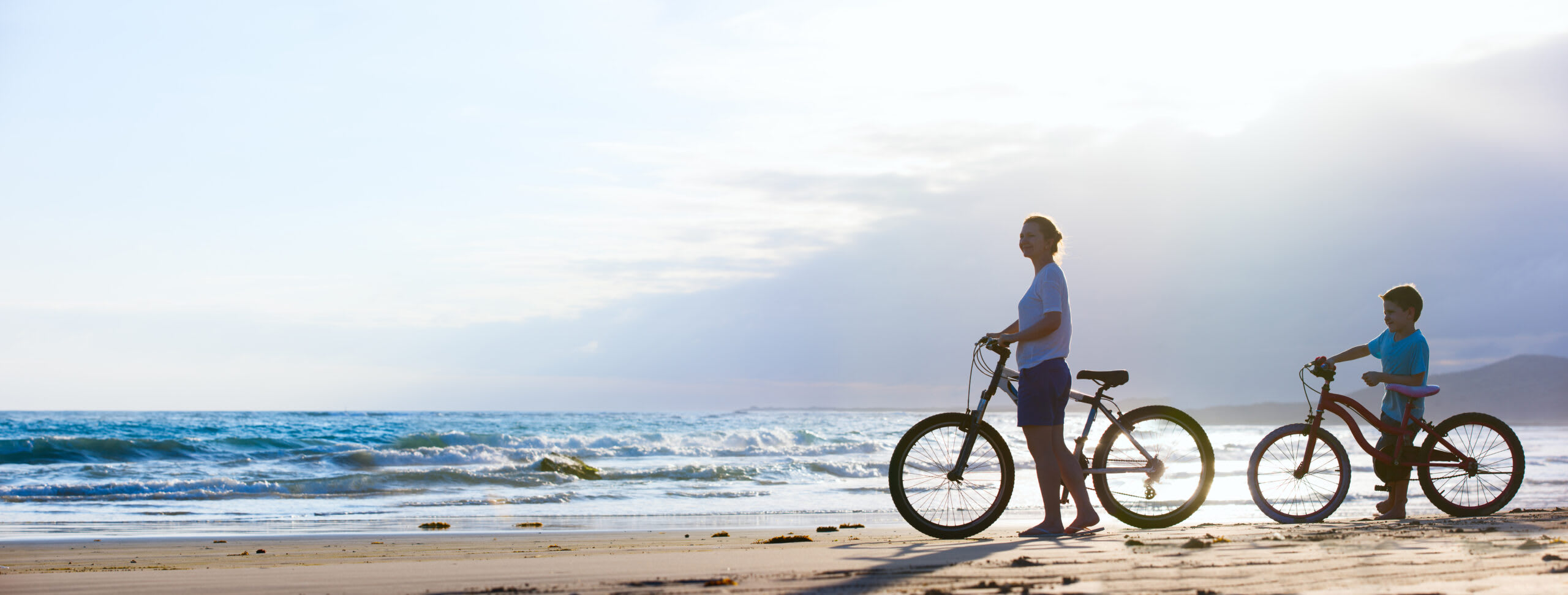 An adult and child ride bicycles along a scenic sandy beach with rolling ocean waves in the background during golden hour. The serene coastal landscape features a calm sky with soft clouds, creating an ideal scene for outdoor recreation and family leisure activities.