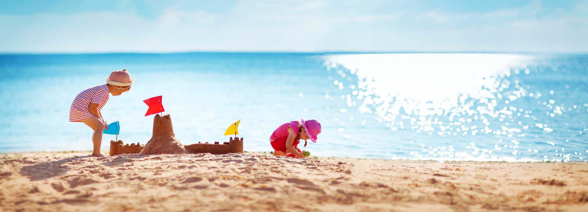 Two young children enjoy a sunny beach day, building an elaborate sandcastle with colorful flags while the sparkling turquoise ocean stretches behind them under clear skies.