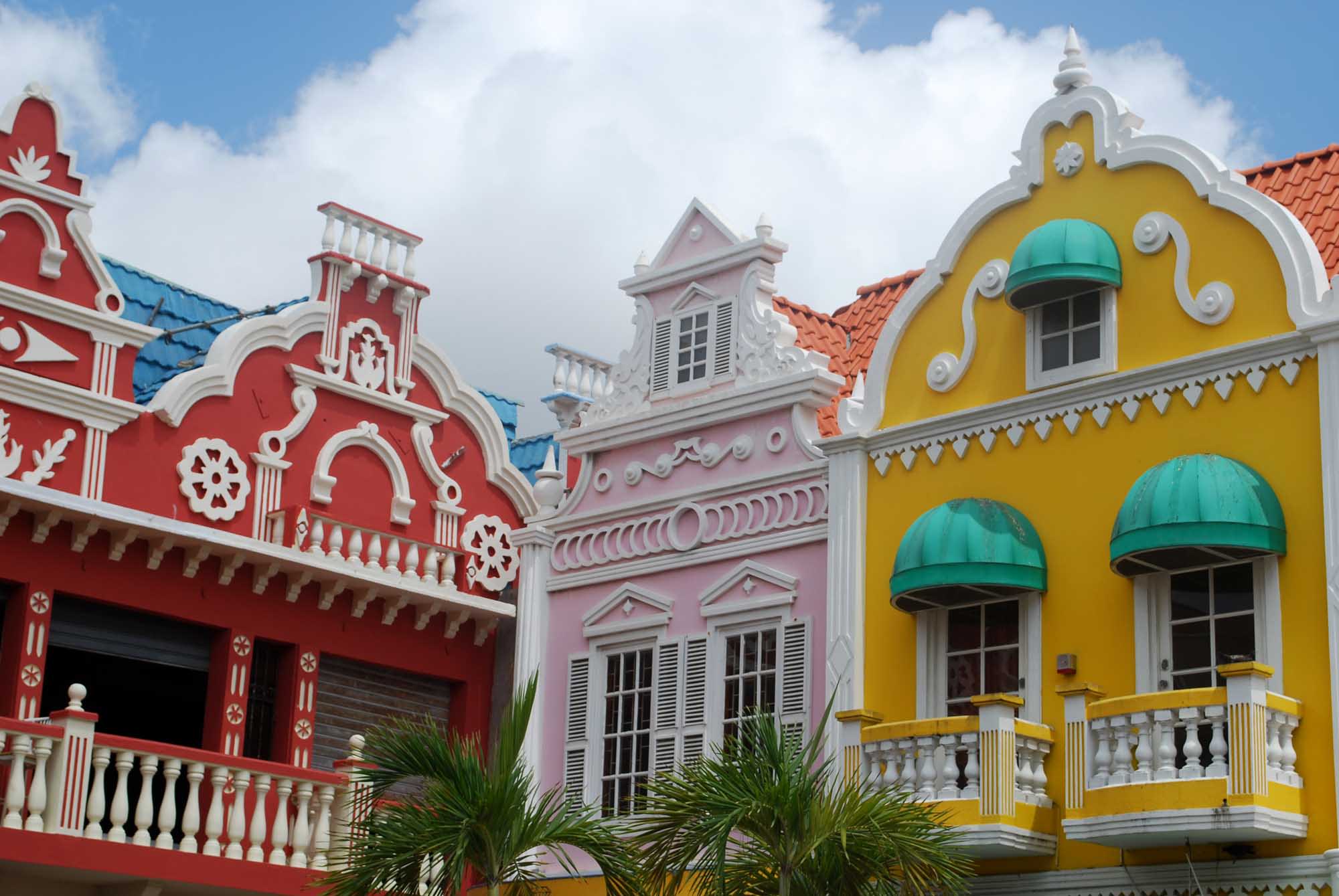 A vibrant row of Dutch Colonial buildings featuring bold primary colors - red, pink, and yellow - with white ornamental trim, arched windows, and turquoise awnings. The iconic architecture reflects Caribbean colonial heritage with distinctive peaked roofs and decorative balustrades, typical of Curacao's historic waterfront district.