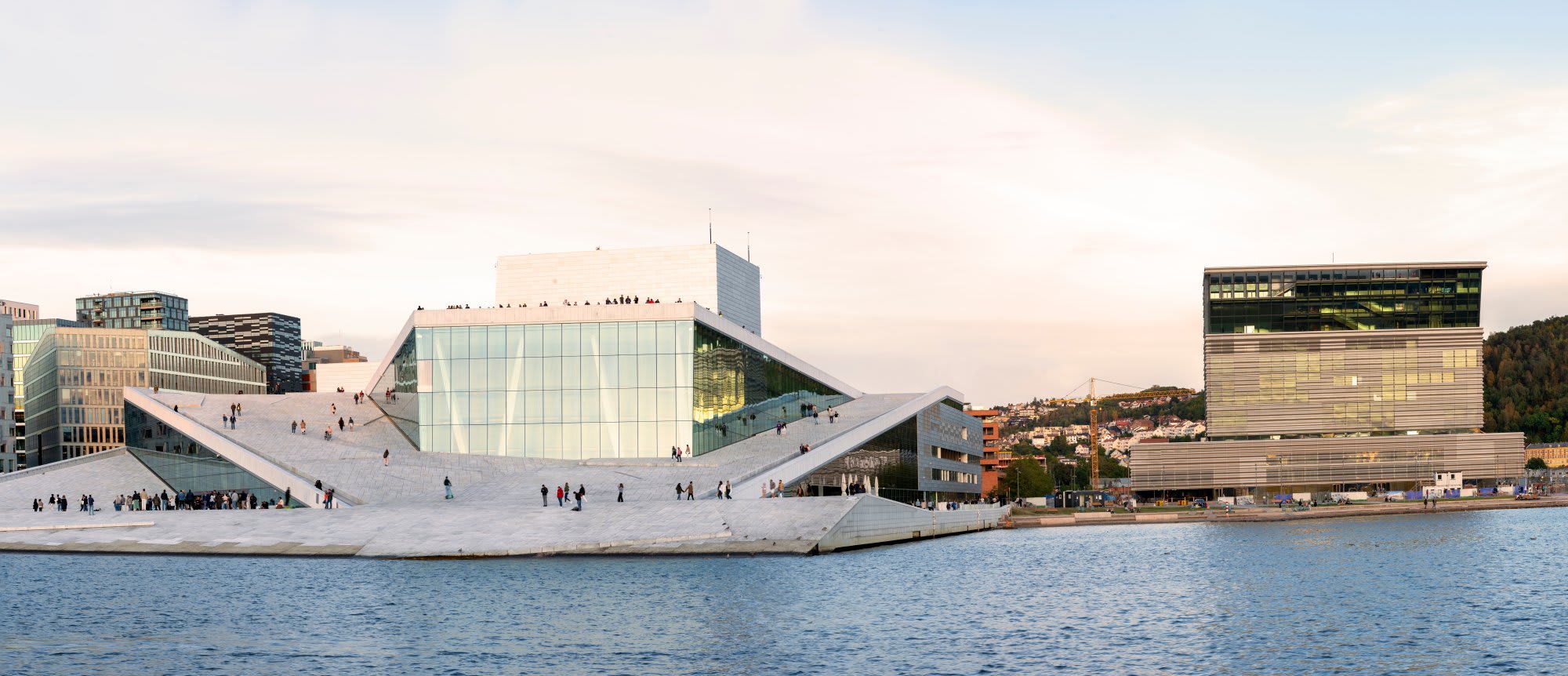 The striking modern Copenhagen Opera House dominates the waterfront with its distinctive white marble sloped architecture and glass facade, flanked by contemporary buildings and reflecting in the calm harbor waters. The iconic cultural landmark showcases Scandinavian design excellence with its bold geometric forms and public plaza.