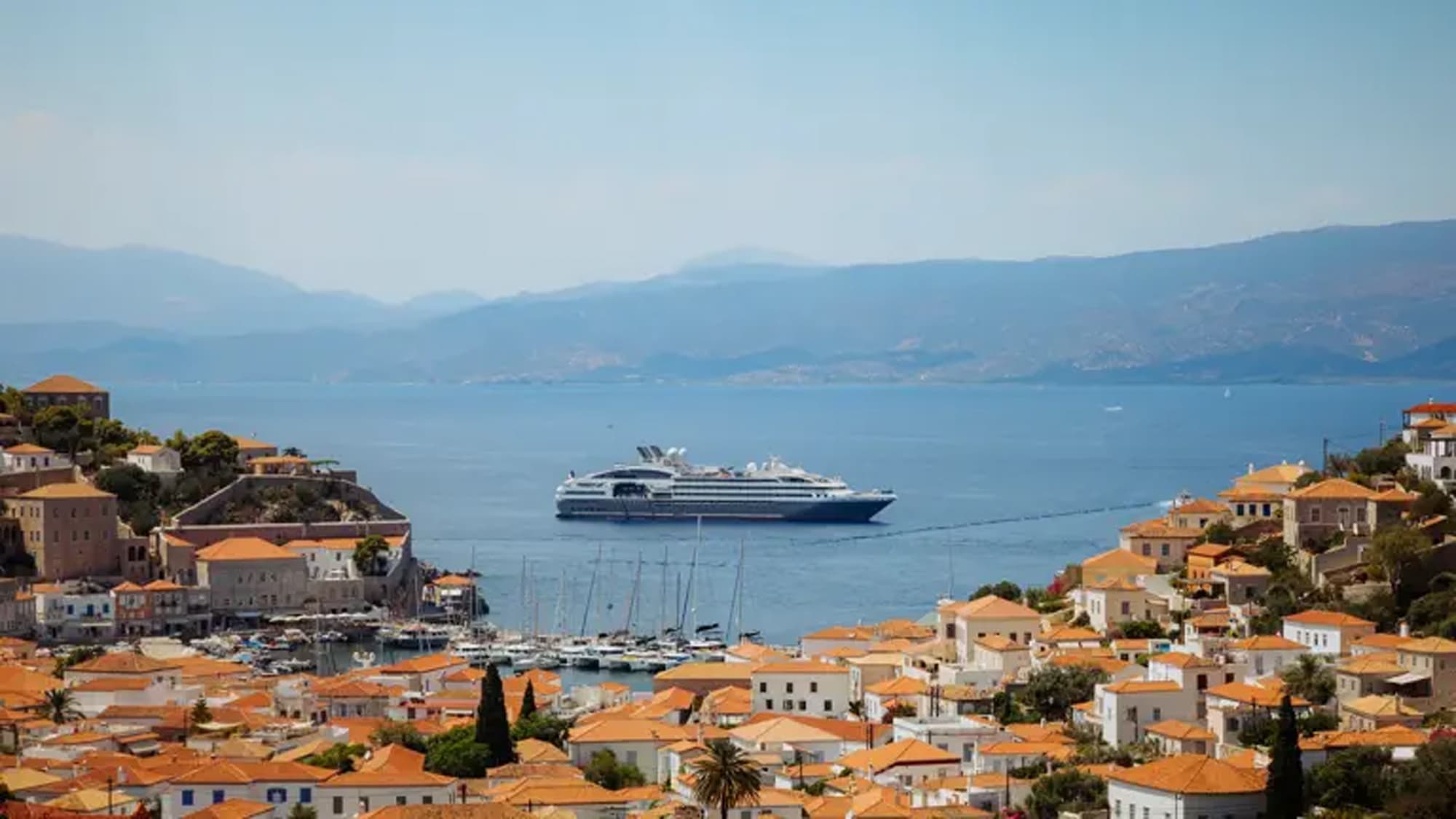 A large modern cruise ship anchored in the crystalline Mediterranean waters off the picturesque Greek island of Hydra, with traditional whitewashed buildings featuring orange-tiled roofs cascading down the hillside.