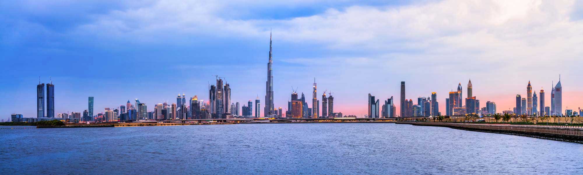 A panoramic view of Dubai's iconic skyline featuring the Burj Khalifa and modern skyscrapers reflected in the calm waters of Dubai Creek during golden hour, with palm trees lining the waterfront.