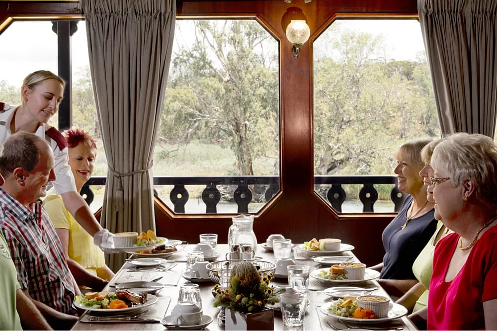 Formal Dining Room with Forest Views An elegant formal dining room aboard a cruise ship with passengers enjoying a meal at a long table. Large windows frame scenic forest and landscape views, while gray curtains, wooden paneling, and warm lighting create a sophisticated atmosphere.