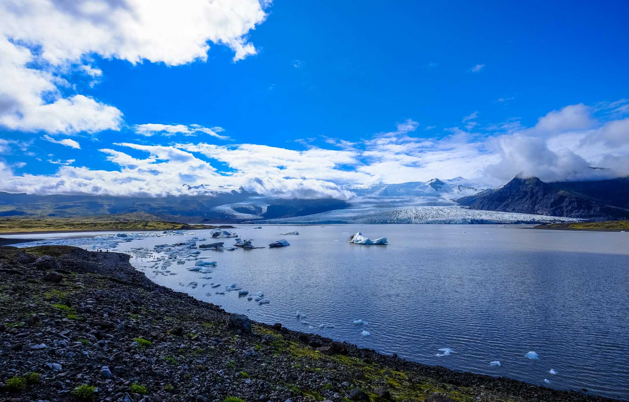 A stunning glacial landscape featuring a pristine blue lake dotted with floating icebergs, with a massive glacier visible in the distance against snow-capped mountains and a bright blue sky. The foreground shows dark volcanic rock shoreline with patches of moss, typical of Iceland's dramatic natural scenery.
