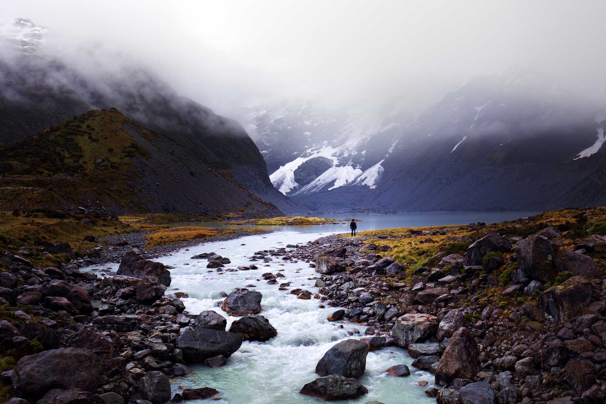 Hiker in Arctic Mountain Valley with Glacier A solitary hiker stands in a remote Arctic valley surrounded by snow-capped mountains shrouded in mist, with a pristine glacial stream flowing across rocky terrain in the foreground. The dramatic landscape features towering peaks with waterfalls and glaciers visible through the fog, creating a stark and otherworldly wilderness scene.