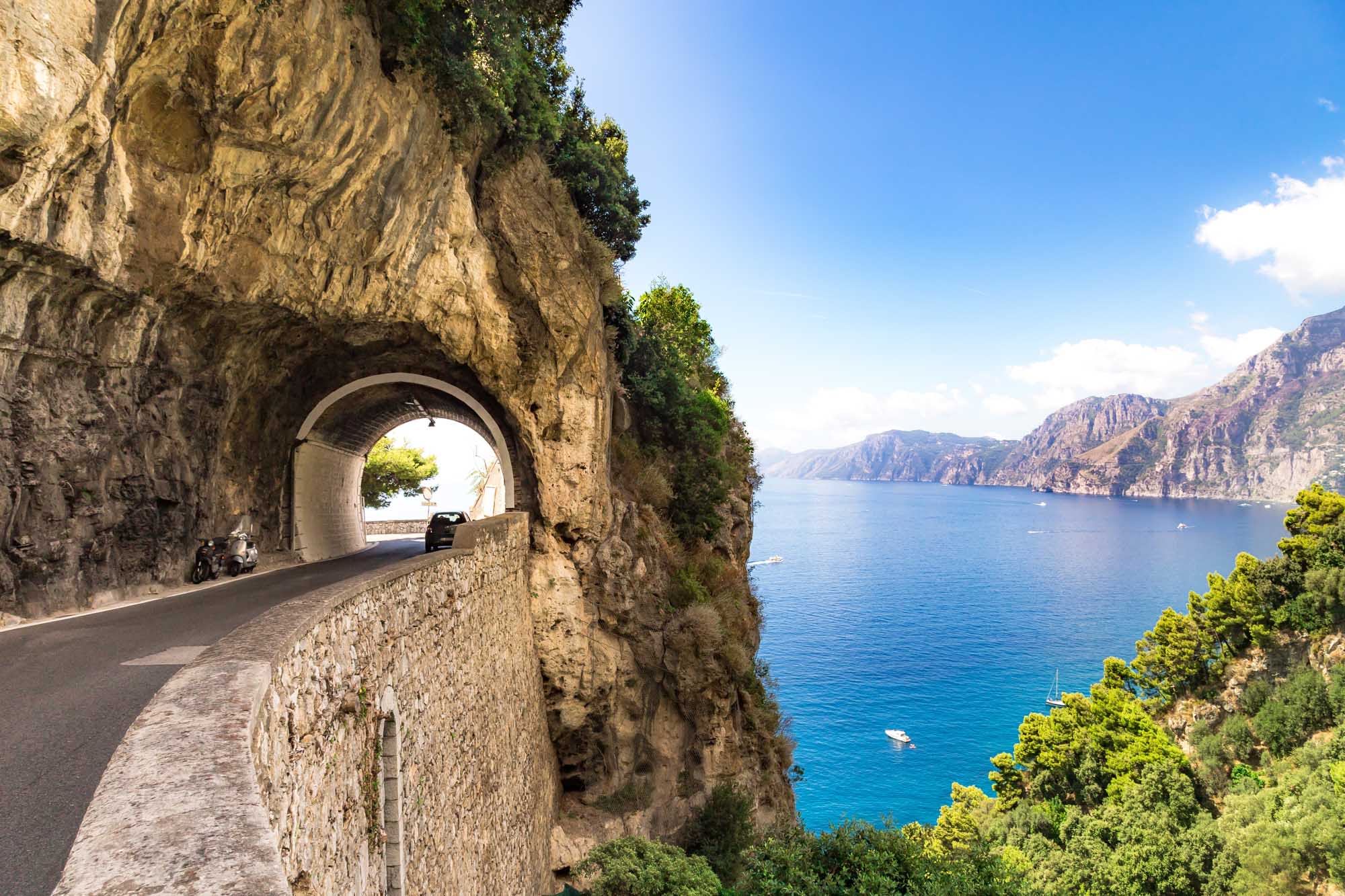 A scenic coastal road carved through a rocky cliff face features a stone tunnel with a scenic Mediterranean vista visible through its arch. The turquoise waters, dramatic cliff formations, and lush Mediterranean vegetation create a stunning backdrop along Italy's famous Amalfi Coast.