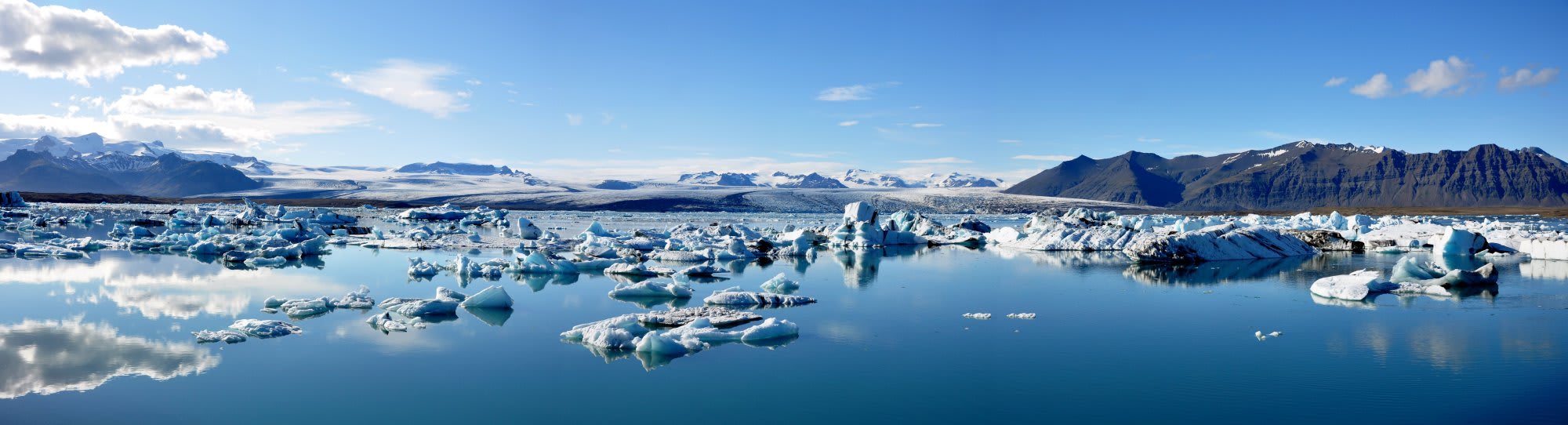 Jökulsárlón Glacier Lagoon Iceland Icebergs A breathtaking panoramic view of Jokulsarlon Glacier Lagoon in Iceland, featuring countless blue and white icebergs floating in pristine turquoise waters. Snow-capped mountains frame the horizon under a clear blue sky with scattered white clouds, showcasing one of Iceland's most stunning natural wonders.
