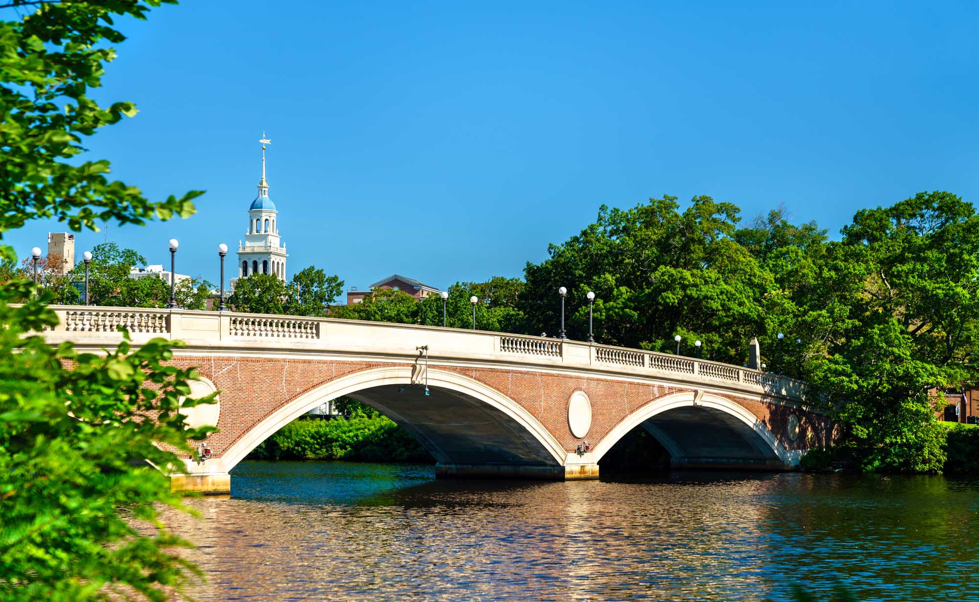 John W. Weeks Memorial Footbridge, Boston The iconic John W. Weeks Memorial Footbridge spans the Charles River between Boston and Cambridge, Massachusetts, featuring distinctive red brick arches and white stone balustrades. A historic church steeple is visible in the background under clear blue skies, with lush green trees framing the scenic riverscape.