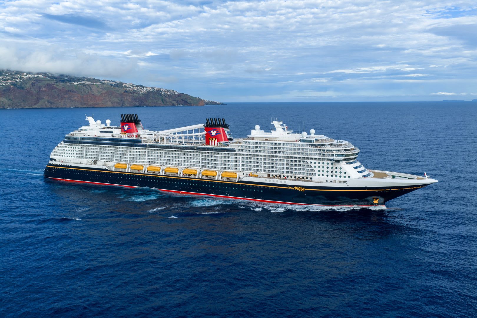 Large Cruise Ship Anchored Off Volcanic Island A massive modern cruise ship with distinctive red funnels featuring Mickey Mouse imagery is anchored in brilliant blue waters off a volcanic island with terraced settlements.